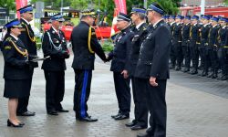 plac, w tle w szeregu stoją strażacy, pośrodku komendant główny gratuluje trzem strażakom